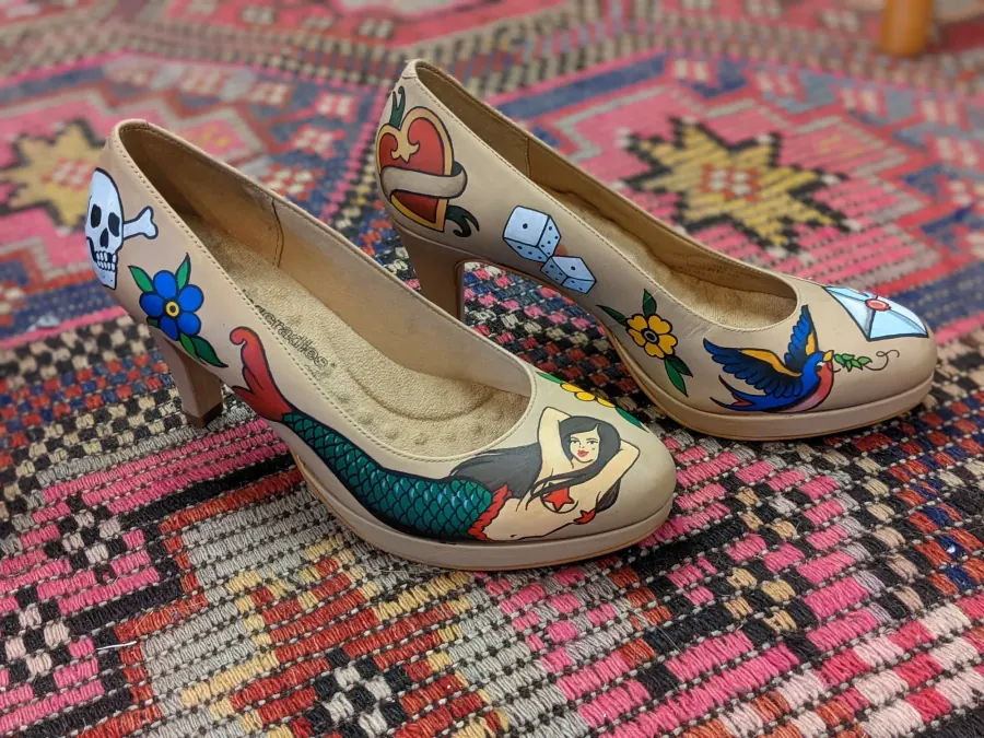 A pair of beige high-heeled shoes with colorful tattoo-style illustrations, including a skull, a mermaid, a swallow, dice, a heart with a banner, a flower, and an umbrella