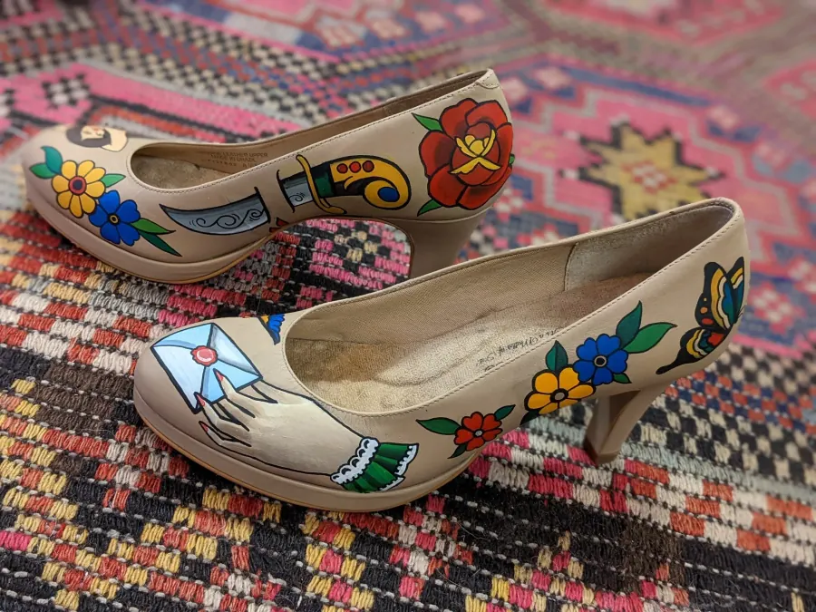 Pair of beige high-heeled shoes with colorful hand-painted floral and tattoo-style designs, placed on a patterned rug