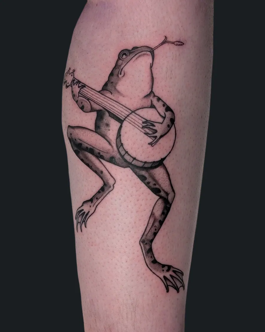 Black and gray tattoo of a frog playing a banjo with a stalk of grass in its mouth by English Cousins, Nashville