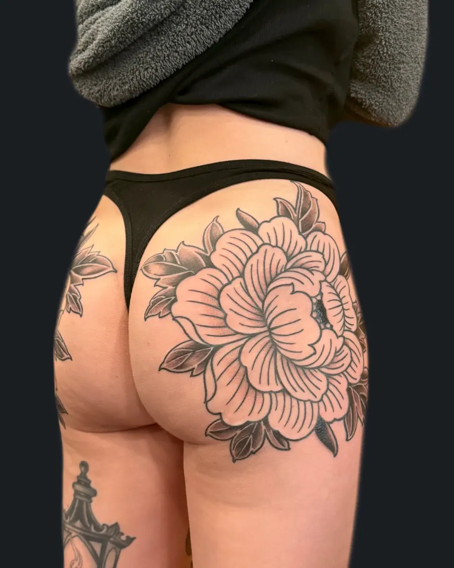 Black and gray floral tattoo featuring a large peony with leaves on the upper thigh by English Cousins, Nashville