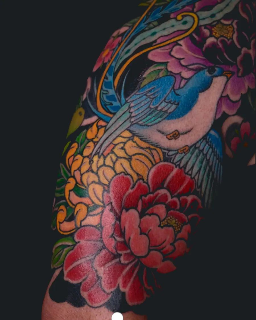 Colorful tattoo of a blue bird flying among large pink and yellow flowers on the arm by English Cousins, Nashville