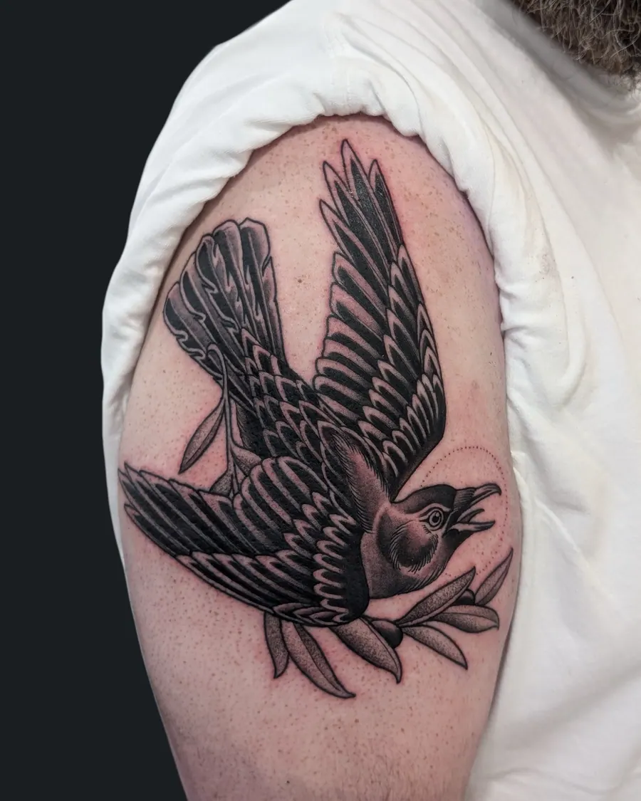 Black and gray tattoo of a crow with outstretched wings carrying an olive branch on the upper arm by English Cousins, Nashville
