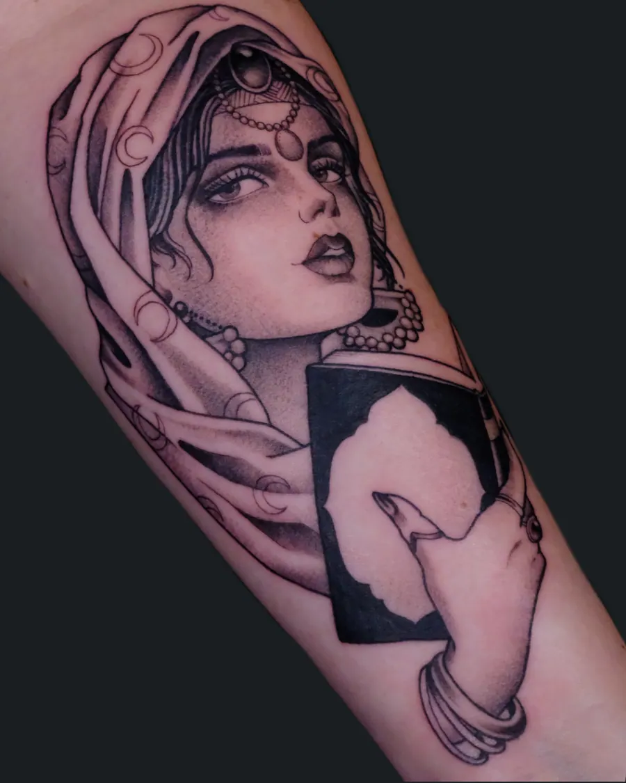 Black and gray tattoo of a woman with jewelry and a crescent-moon head covering holding a card on the inner arm by English Cousins, Nashville