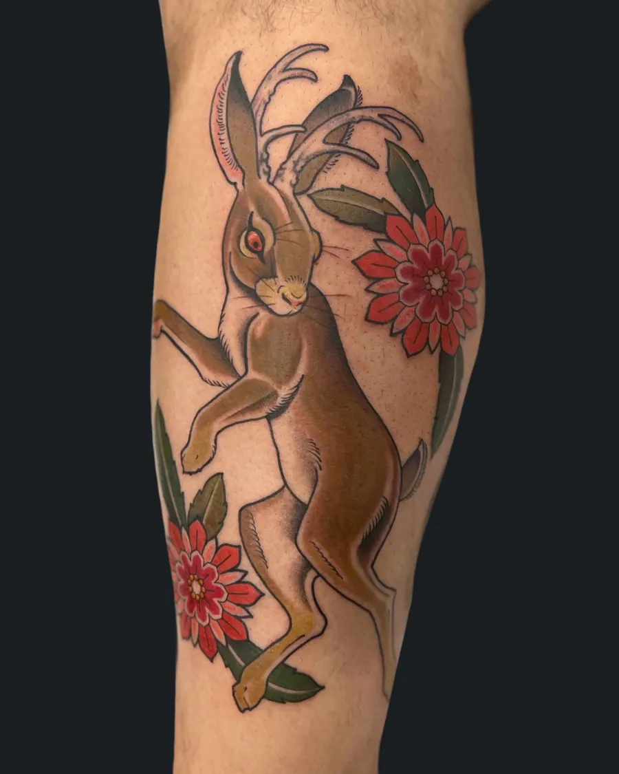 Neotraditional jackalope tattoo with red flowers and leaves on the calf by English Cousins, Nashville