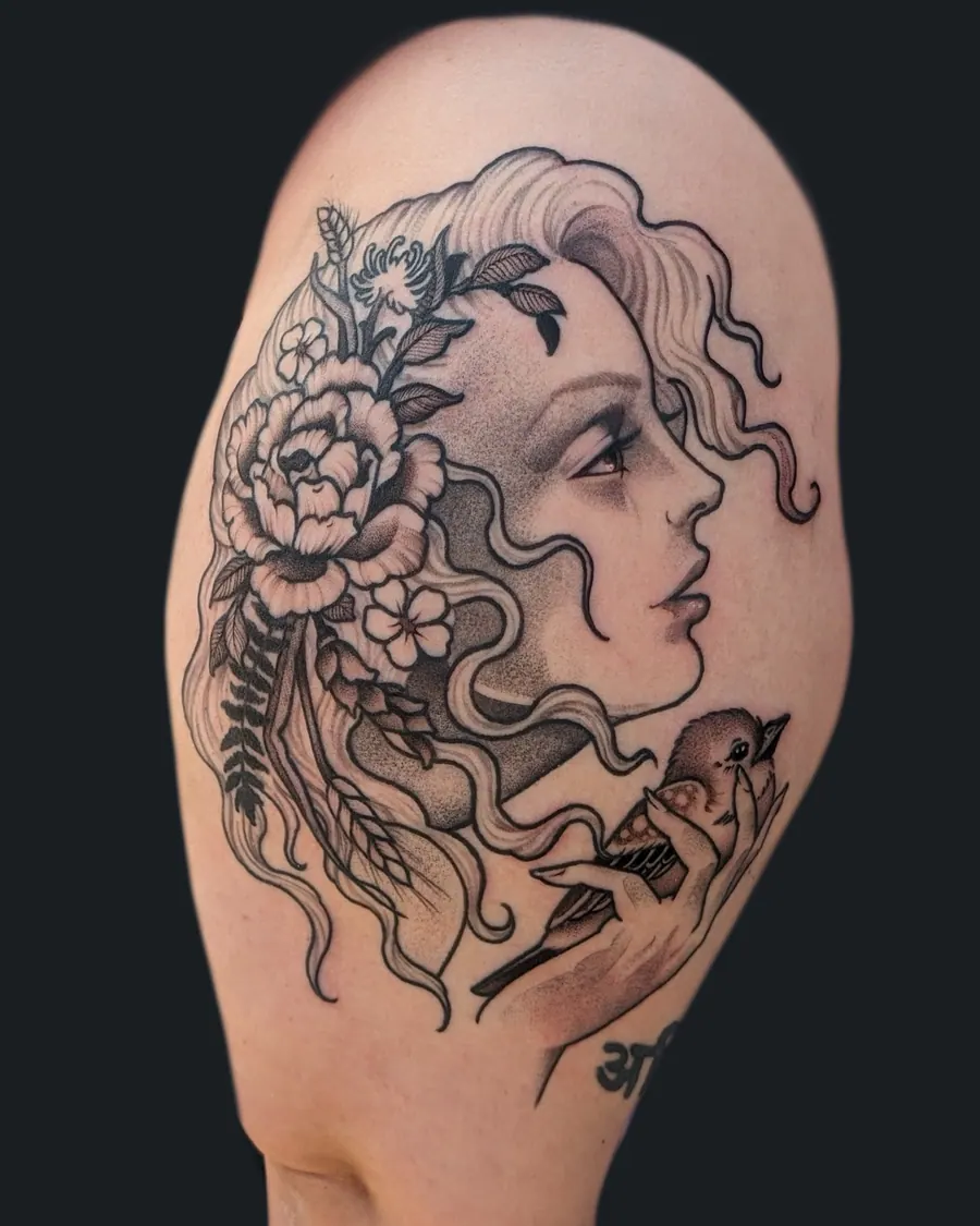 Black and gray tattoo of a woman's profile with flowers in her hair, holding a small bird, on the shoulder by English Cousins, Nashville