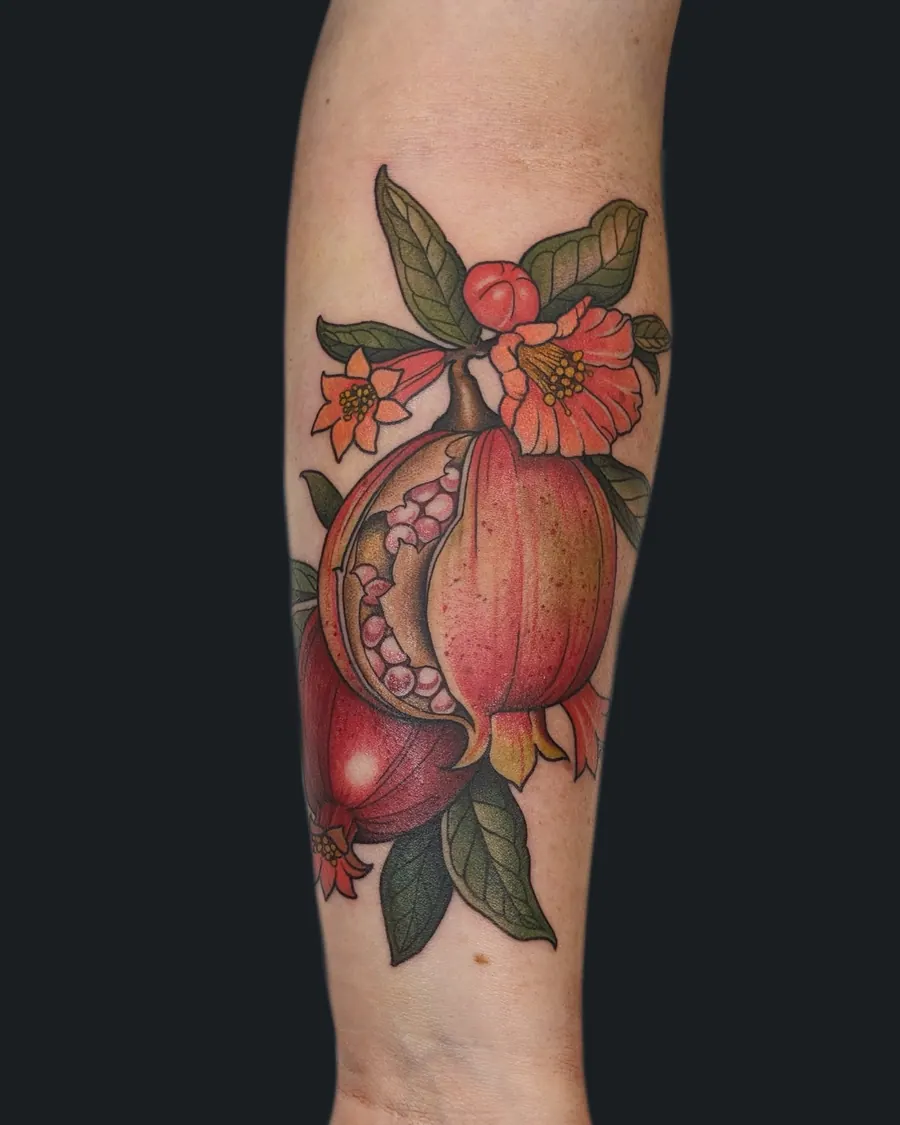 Neotraditional pomegranate tattoo, split to show its seeds, with red blossoms and leaves on the forearm by English Cousins, Nashville