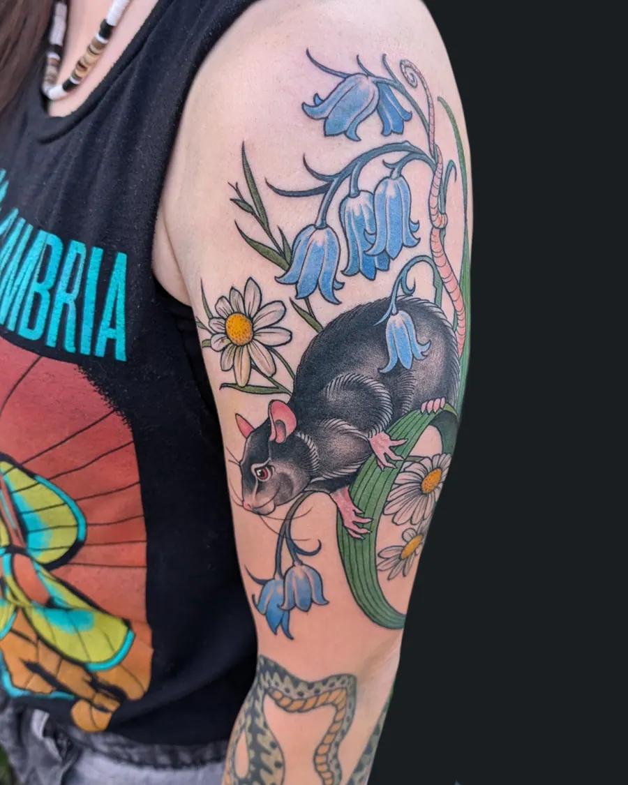Colorful tattoo of a black rat with red eyes surrounded by bluebell flowers and daisies on the upper arm by English Cousins, Nashville